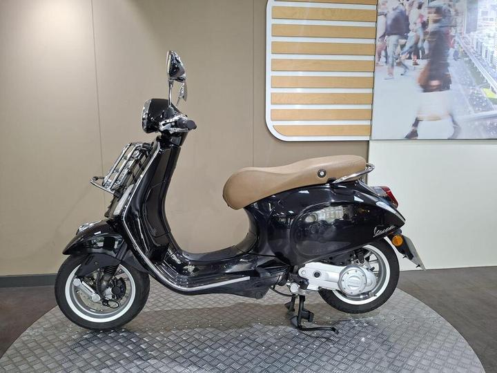 A black Vespa scooter with a brown seat displayed on a circular platform, set against a minimalist background with a large screen showing a bustling urban scene.