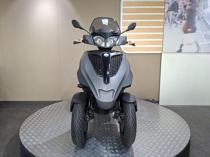 A gray three-wheeled scooter is displayed on a circular platform, facing forward, with its headlights on and a blurred background featuring a bustling urban scene.