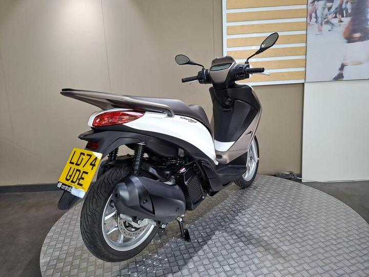 A white and grey scooter displayed on a circular metal platform, showing the rear and side view, with visible license plate "LD74 UDE" and a simple background.
