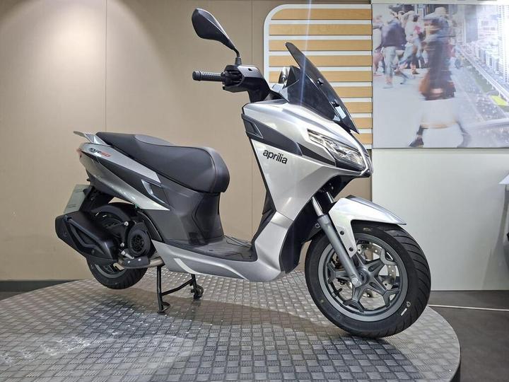 A silver Aprilia scooter displayed on a circular platform, with a neutral background featuring a blurred urban scene.