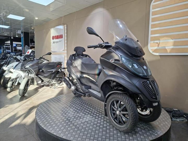 A black three-wheeled scooter displayed on a circular stand inside a showroom, with other scooters in the background and a Yamaha sign on the wall.