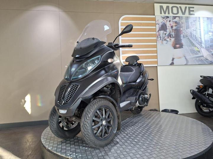 A black three-wheeled scooter displayed on a circular pedestal in a modern showroom, featuring a windscreen and distinct design elements. Behind it, a wall displays the word "MOVE" and an image of pedestrians.