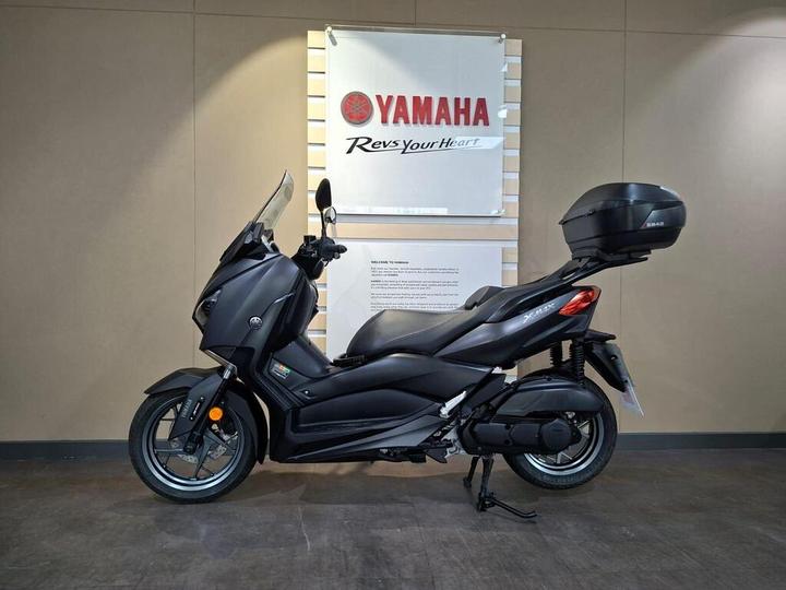 A sleek black Yamaha XMAX scooter displayed in a showroom setting, with a promotional banner in the background highlighting the Yamaha logo and slogan.