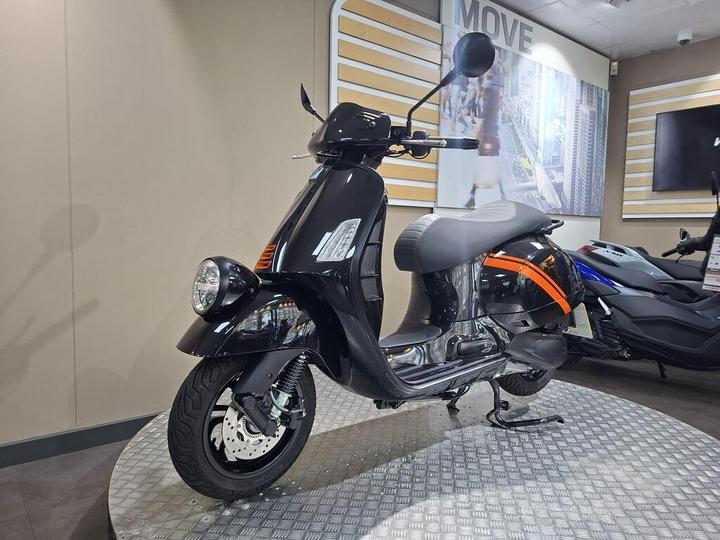 A modern black scooter with a sleek design and an orange stripe, displayed on a metal platform in a showroom against a light-colored wall and promotional graphics.