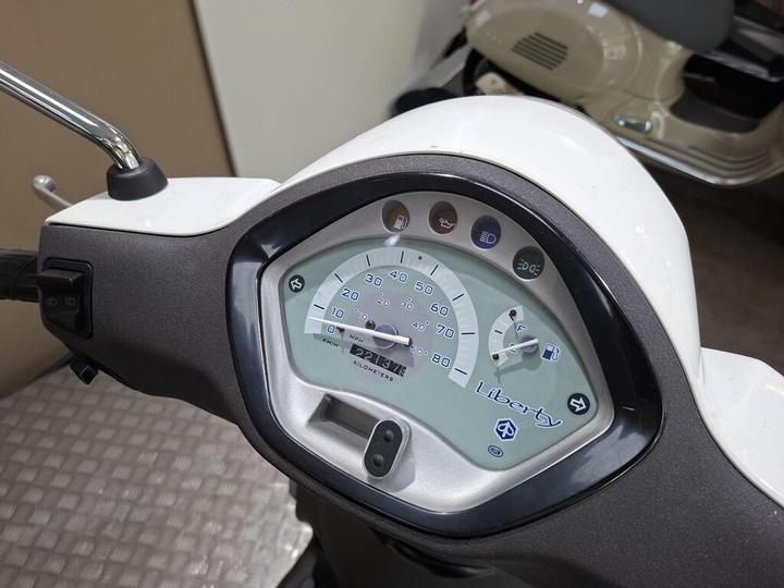 Close-up of a scooter dashboard featuring speedometer and fuel gauge, with indicators for lights and engine status. The design includes a prominent "Liberty" logo.