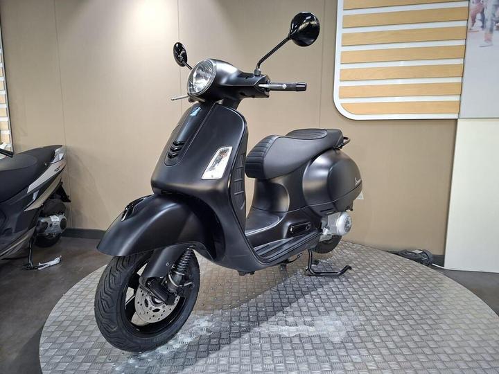 A sleek black scooter displayed on a circular platform, featuring a rounded headlight, a textured seat, and stylish mirrors, with a plain background emphasizing its design.