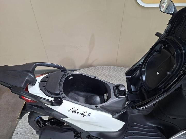 A close-up view of the storage compartment of a white and red scooter with the "Liberty S" logo, showing an open lid and a round fuel cap inside.