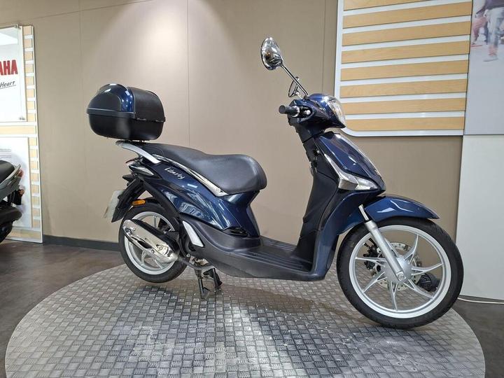 A blue scooter with a black top case is displayed on a textured round platform in a showroom. The background features a neutral-colored wall and Yamaha branding.