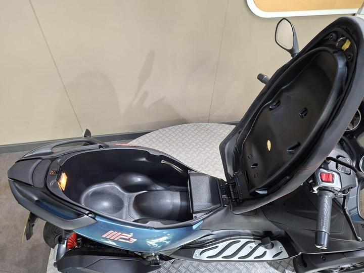A top view of a motorcycle seat with the storage compartment opened, revealing a spacious, black interior. The motorcycle is positioned on a textured surface, and part of the dashboard is visible on the right side.