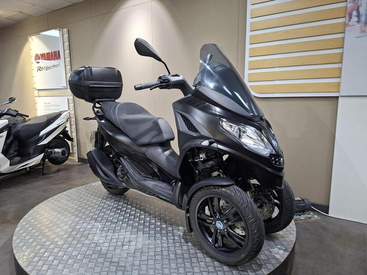 A black three-wheeled scooter with a sleek design, featuring a comfortable seat and a rear storage compartment, displayed on a circular metallic platform in a showroom.