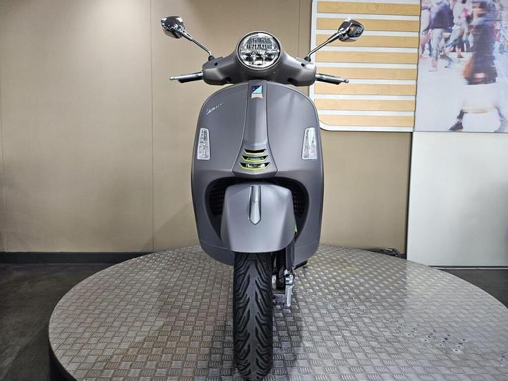 Front view of a gray scooter displayed on a circular platform, with a checkered floor pattern and a blurred background featuring people and a wooden panel.