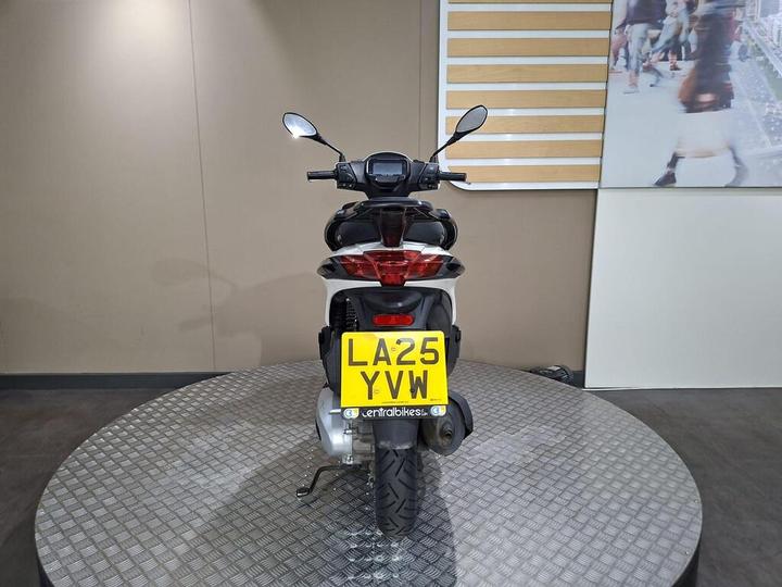 A rear view of a black scooter displayed on a circular platform, featuring a visible license plate with the number LA25 YVW. The background is a neutral-colored wall with a partially visible graphic panel.