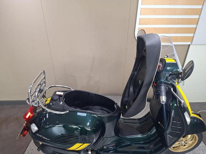 A green scooter with yellow stripes featuring a sidecar, displayed in a showroom with neutral-colored walls. The sidecar is empty and visibly spacious.