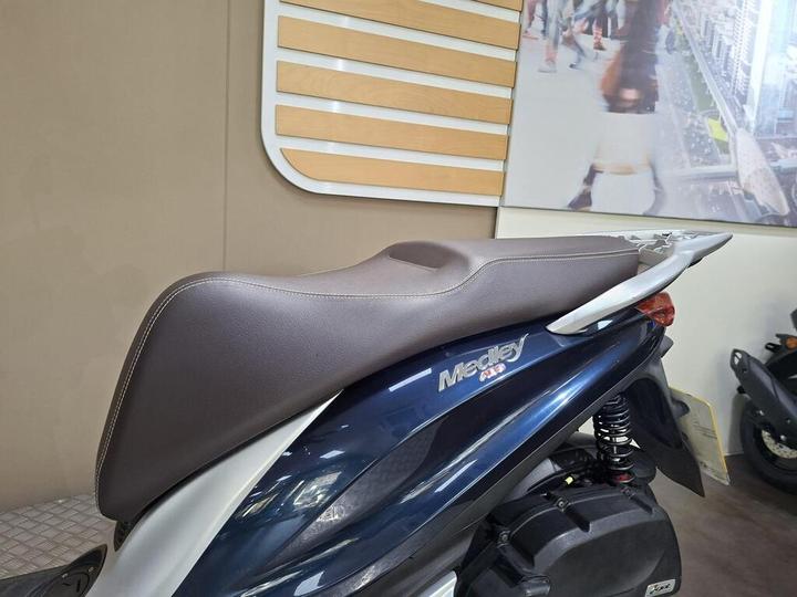 A close-up view of a scooter's seat and rear section, featuring a dark blue color and a brown seat upholstery with white stitching, displayed in a showroom setting.