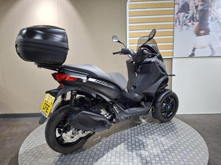 A sleek black three-wheeled scooter with a rear top box, displayed on a patterned circular platform against a neutral wall.