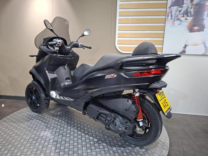 A sleek black scooter with a large windshield and comfortable seating, showcased on a textured circular platform against a neutral background.