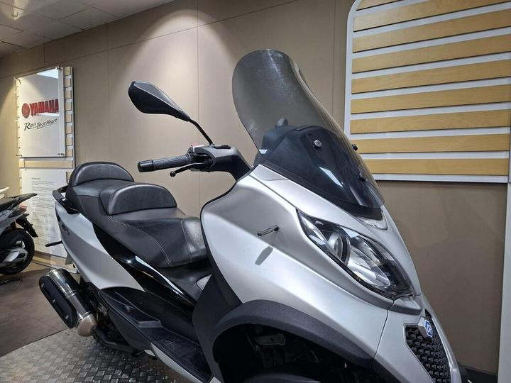 A silver scooter with a windshield is displayed in a showroom, featuring black seating and handlebars, against a neutral wall with Yamaha branding.