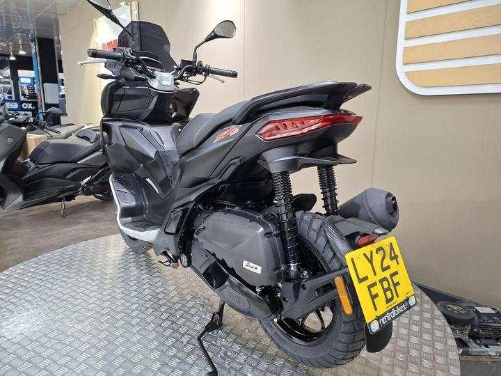 A black scooter displayed at a showroom, featuring a sleek design, prominent rear tail light, and a visible license plate reading "LY24 FBF." The scooter is positioned on a textured surface with other scooters faintly visible in the background.