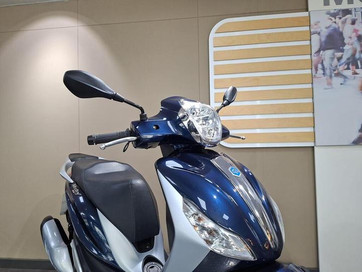 A close-up view of a blue and silver scooter with a prominent headlight, handlebars, and mirrors, set against a neutral wall background featuring wooden accents.