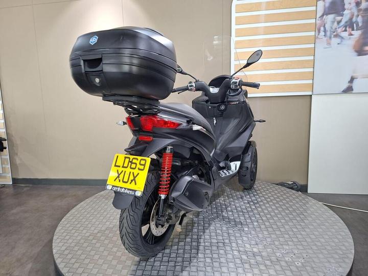 A black scooter displayed on a rotating stand, featuring a top case and a clear windshield, with a license plate reading "LD69 XUX." The background is minimalistic, with neutral colors and a soft texture.