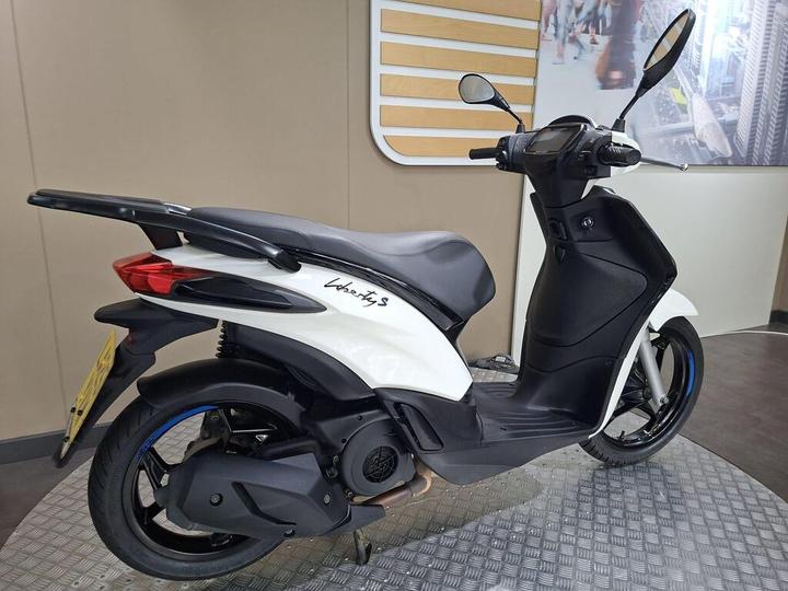 A white scooter with a sleek design, featuring a black seat and rear tail light, positioned on a textured floor against a plain wall backdrop.