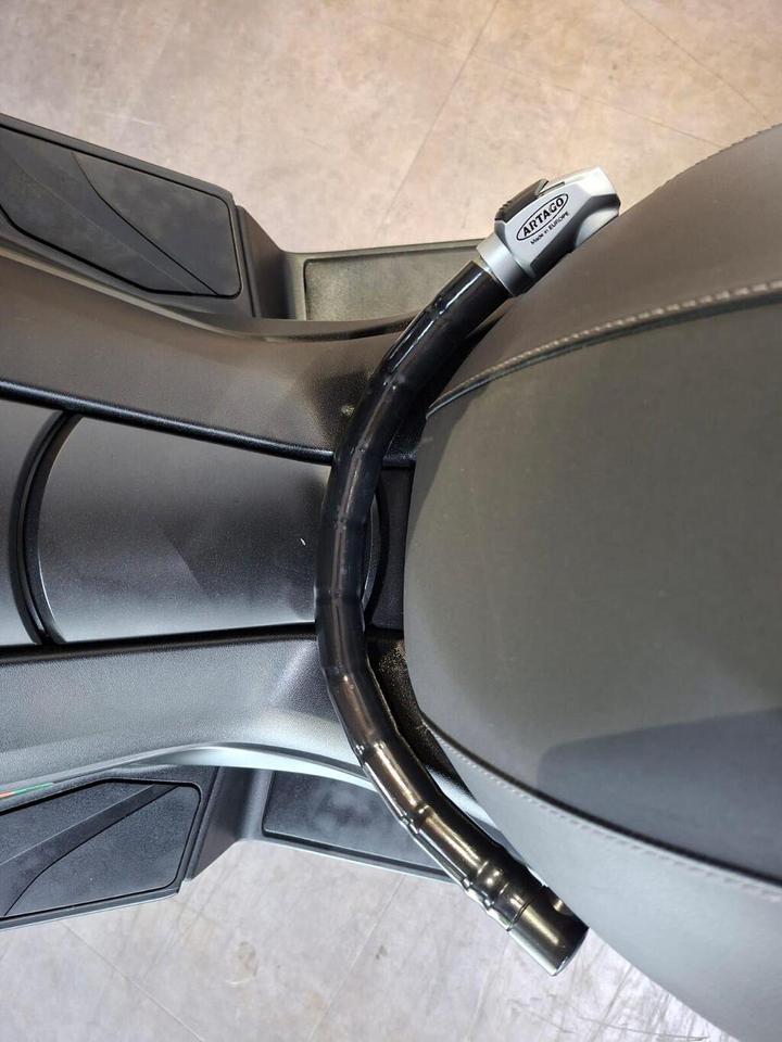 A close-up view of a motorcycle seat and lock, featuring a black cable lock secured around the seat, with a silver lock mechanism visible on the right. The background shows a textured floor surface.