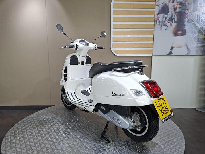 A white Vespa scooter is displayed in a showroom, facing slightly to the right with its back visible. The scooter features a sleek design, chrome accents, and a license plate reading "LG71 XSA." The background includes a light-colored wall and a blurred image of people walking.