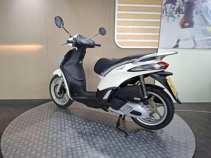 A white and black scooter parked on a circular metallic platform, shown from the rear left side, with a minimalist background.