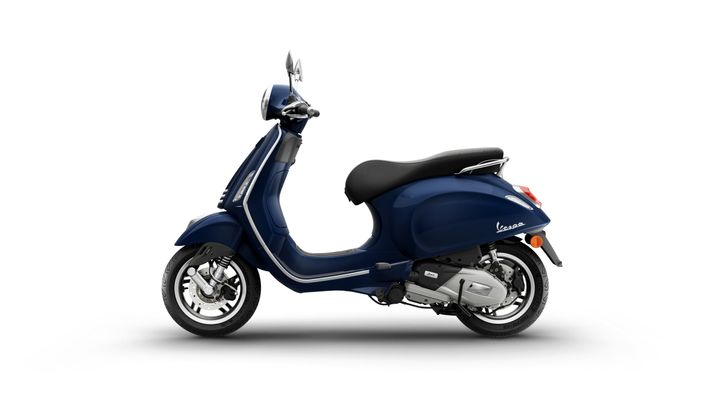 A blue Vespa scooter viewed from the side, showcasing its sleek design and chrome accents against a plain white background.