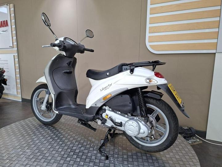 A white scooter parked on a textured floor, showcasing its sleek design, rear view, and prominent features like mirrors and a seat. The background includes a minimalist wall with wooden accents.