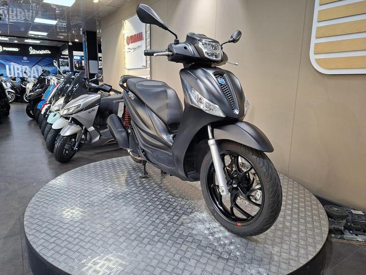 A sleek, matte black scooter displayed on a circular metallic platform in a showroom, surrounded by various other scooters in the background.