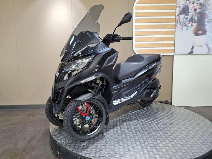 A sleek black three-wheeled scooter with a tinted windscreen, displayed on a circular platform with a textured surface. The scooter features modern design elements, including sporty wheels and a streamlined body. Background includes a large graphic with blurred people.