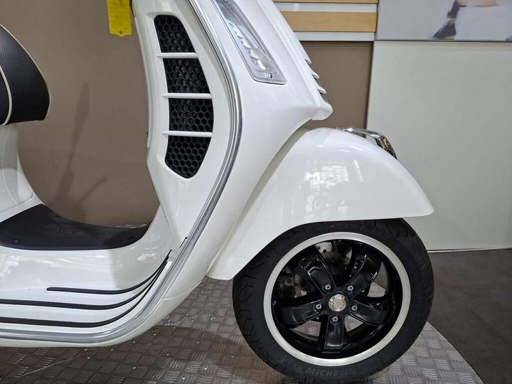 A close-up view of the front right section of a white scooter, showcasing a sleek design, a black wheel with silver accents, and a portion of the scooter's body with modern lines and details.