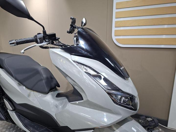 A close-up view of a silver Honda scooter, showcasing the handlebars, seat, and aerodynamic front design with a dark windshield. The background features a neutral-toned wall with wooden panel accents.