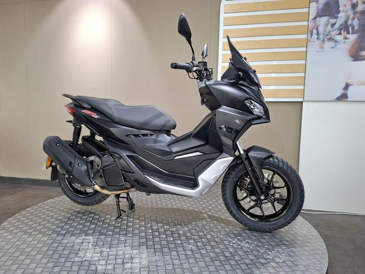A sleek black and silver scooter is displayed on a textured circular platform, with a neutral background showcasing a partially visible mural of people. The scooter features a modern design with a prominent front windshield and dual headlights.