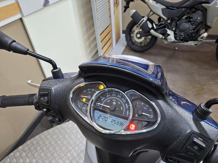 Close-up view of a scooter's dashboard displaying gauges for speed and fuel, with indicators lit up and a digital clock showing 2:20. A motorcycle is visible in the background.