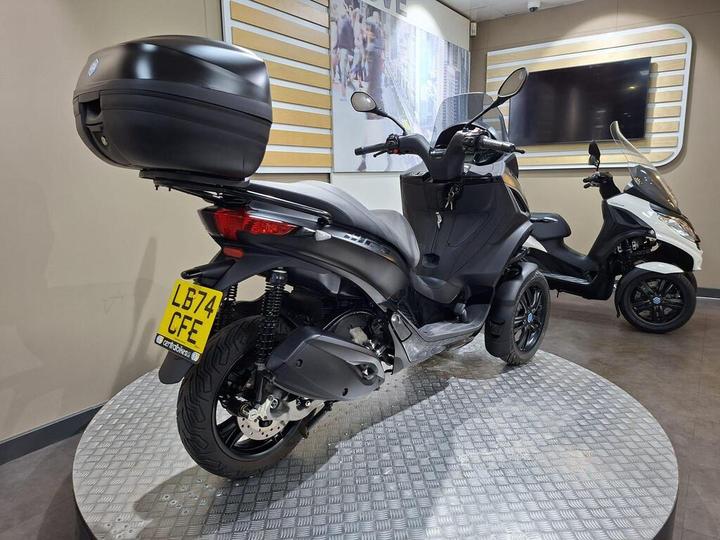 A black three-wheeled scooter with a top case is displayed on a circular platform, alongside a white scooter in a modern showroom setting.