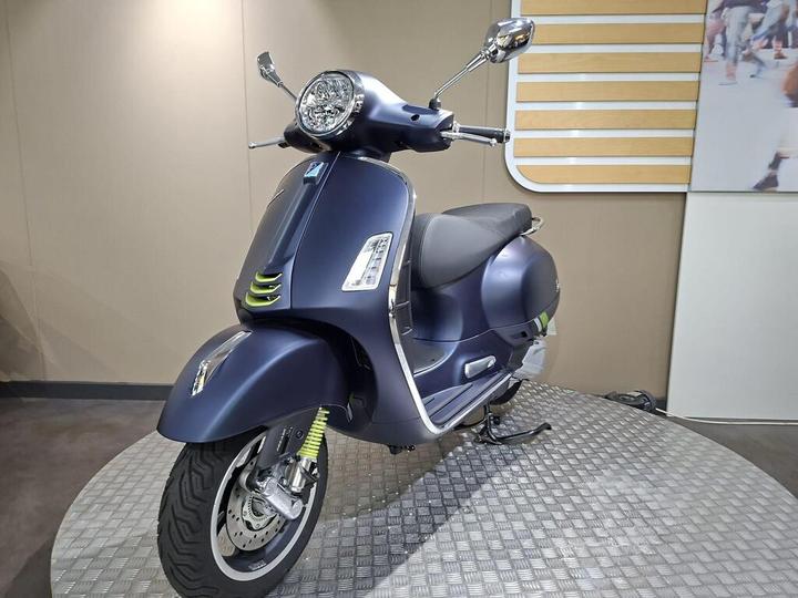 A dark blue scooter displayed on a textured platform, featuring a round headlight, chrome accents, and a sleek design with green highlights.
