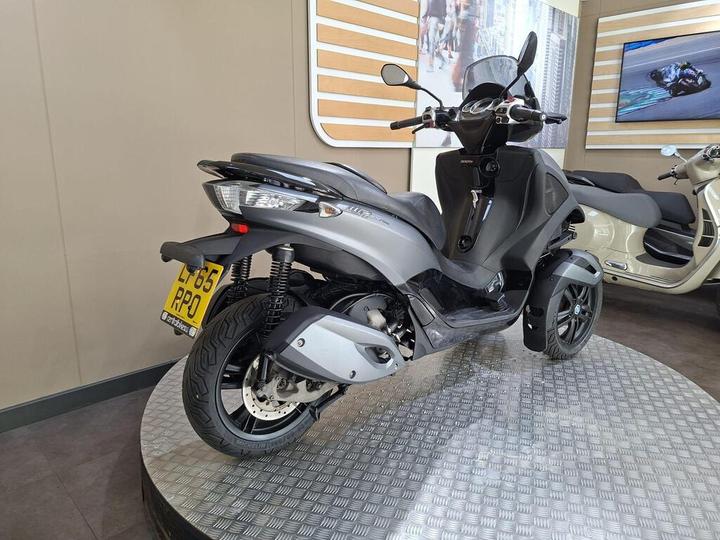A gray scooter displayed in a showroom, featuring two front wheels and a stylish design, with a backdrop of a wall and a screen showing motorcycle-related content.