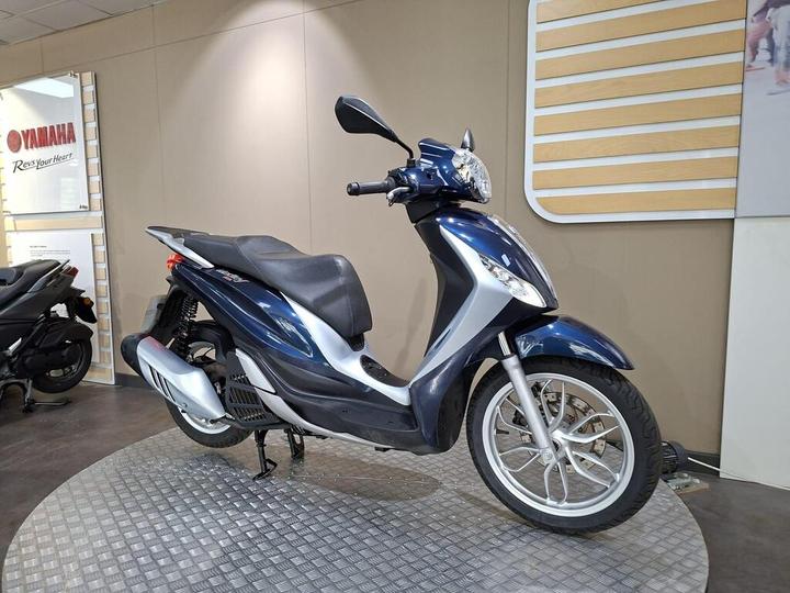 A blue and silver scooter is displayed on a round metal platform in a showroom with a Yamaha logo in the background.