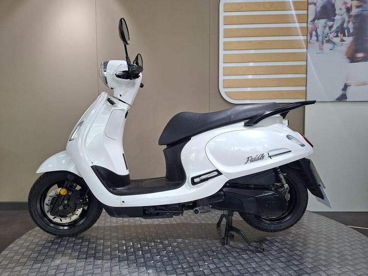 A white scooter is displayed in profile on a circular textured platform, with a minimalist background featuring neutral tones and a blurred image of people in motion.