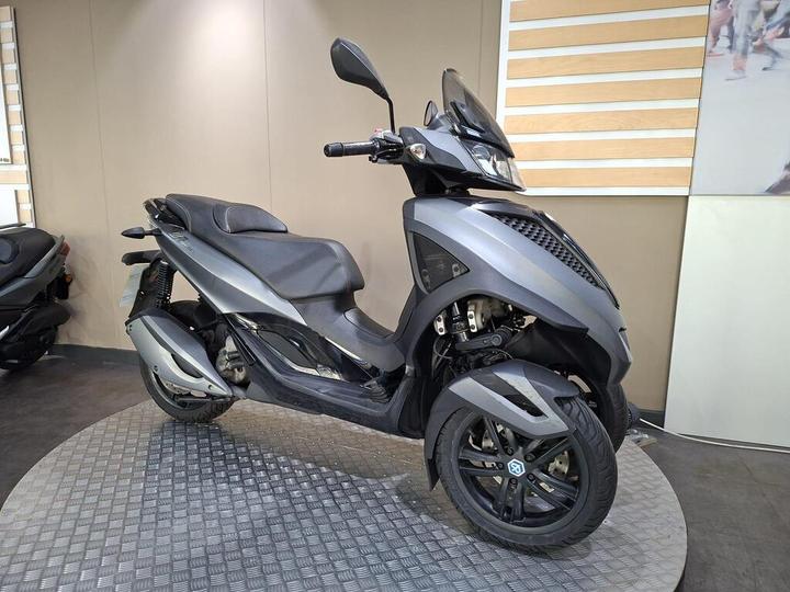 A modern three-wheeled scooter with a sleek gray design is displayed on a circular platform in a showroom, featuring a comfortable seat and a clear windshield.