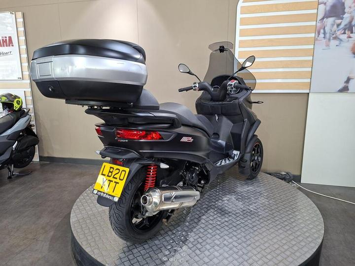 A black three-wheeled scooter with a silver top case stands on a circular display platform in a showroom. The background features a neutral wall and a promotional sign. The scooter has red suspension components and a license plate visible.