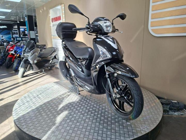 A black scooter displayed on a rotating platform in a motorcycle showroom, with various other motorcycles visible in the background.