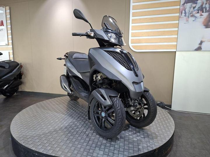 A modern grey scooter with three wheels displayed on a raised platform in a sleek showroom setting.
