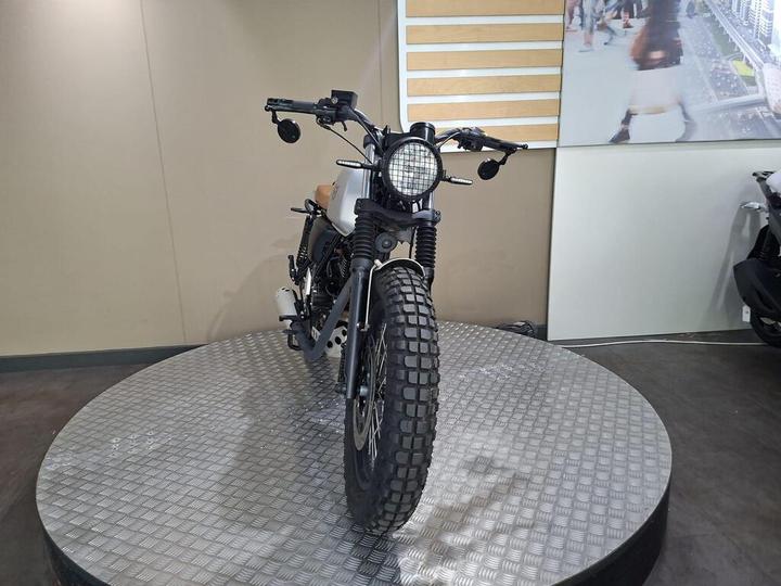 A motorcycle displayed on a circular platform, viewed from the front with its distinctive round headlight and rugged tires, set against a neutral back wall in a showroom.