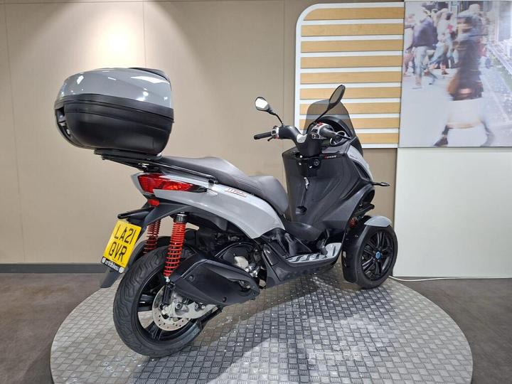 A sleek gray three-wheeled scooter with a top case parked on a circular metal platform, set against a modern interior backdrop.
