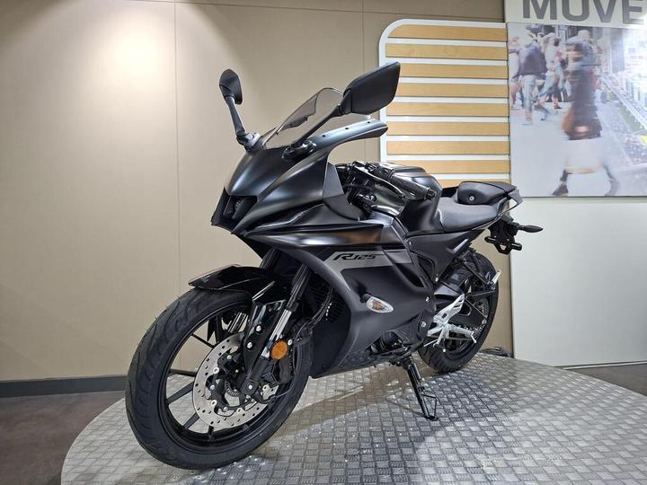 A sleek black motorcycle, the Yamaha R125, is displayed on a circular platform with a textured surface. The background features a light-colored wall with graphic elements, while the motorcycle is angled to showcase its aerodynamic design and modern features.