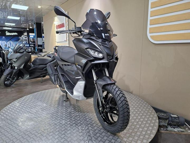 A sleek black motorcycle displayed on a circular metal platform, showcasing its prominent front view, with a windscreen and distinctive headlight design. Other motorcycles are partially visible in the background, along with a branded display wall.