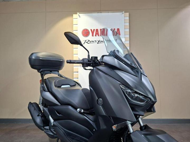 A sleek, black Yamaha scooter is shown from the front-right angle, featuring a tall windshield and a top box, with a logo backdrop reading "Yamaha, Rev Your Heart."